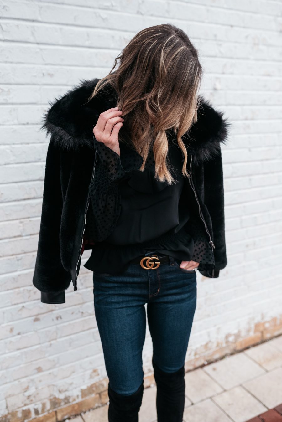 Suzanne wearing a black blouse, faux fur jacket, denim, belt and over the knee boots