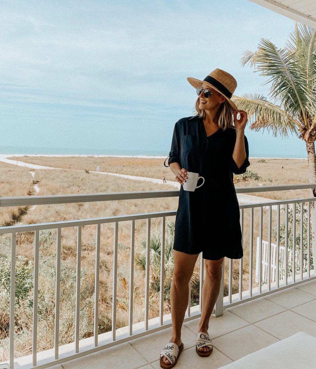 Suzanne wearing a swimsuit cover up, straw hat, sandals and sunglasses while holding a cup of coffee on the balcony overlooking the beach 