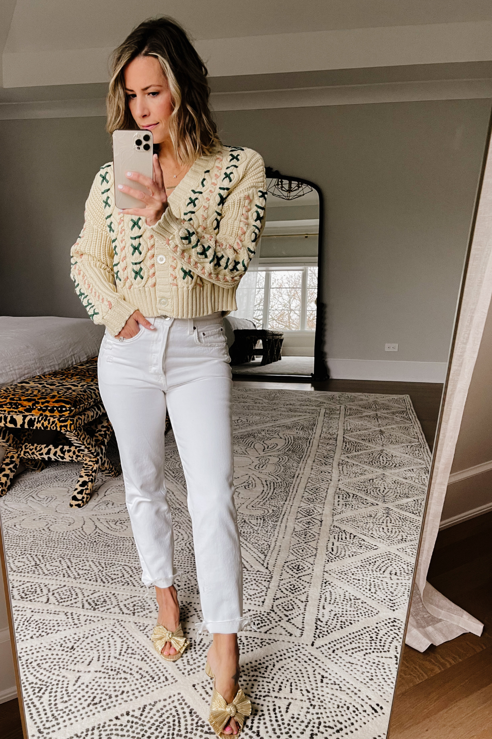 Suzanne taking a mirror selfie wearing a cable cardigan, white cropped denim, and gold metallic heels.