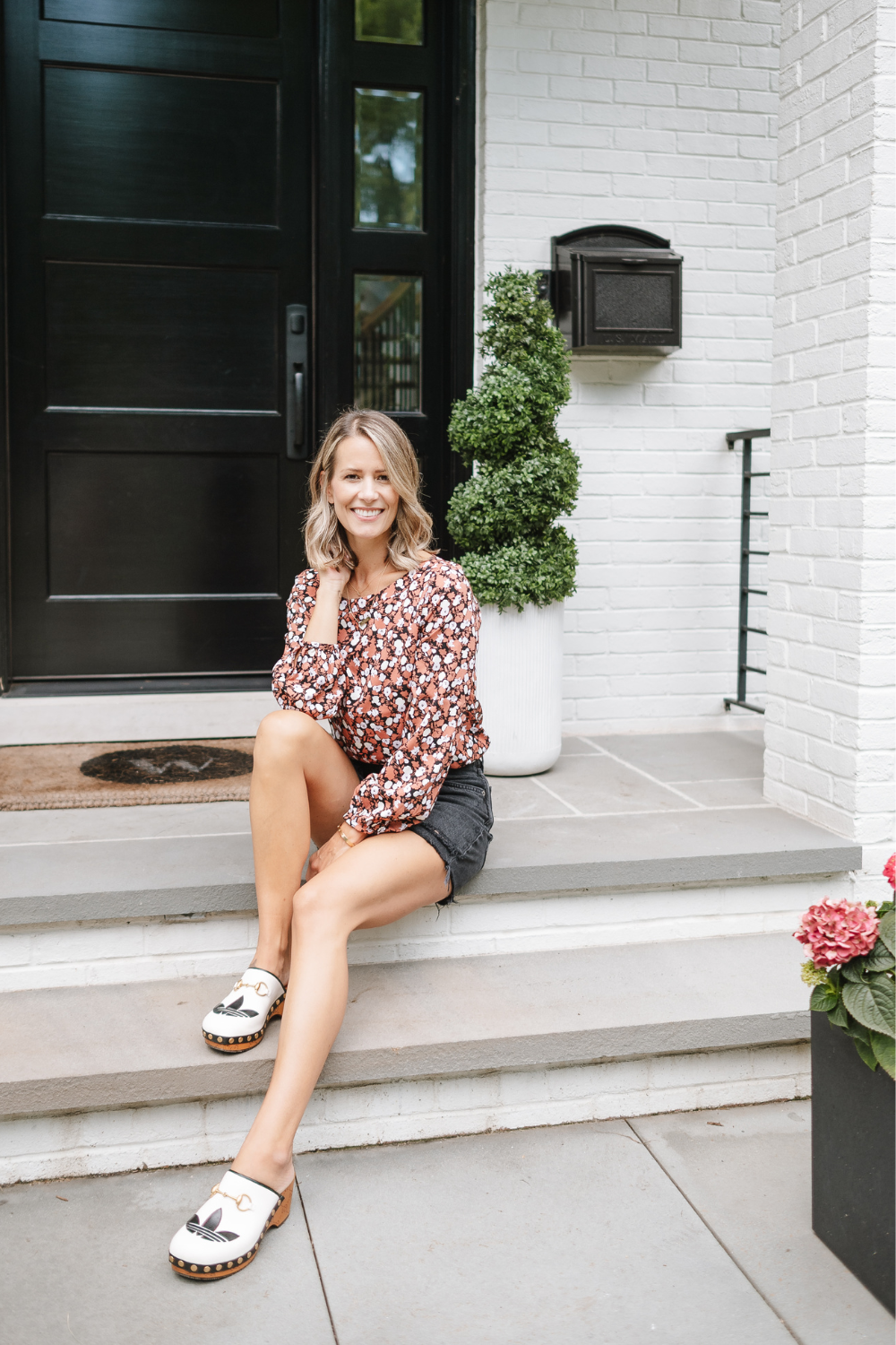 Suzanne on her front porch wearing a low price floral Amazon top, black denim shorts, and designer Gucci X Adidas clogs