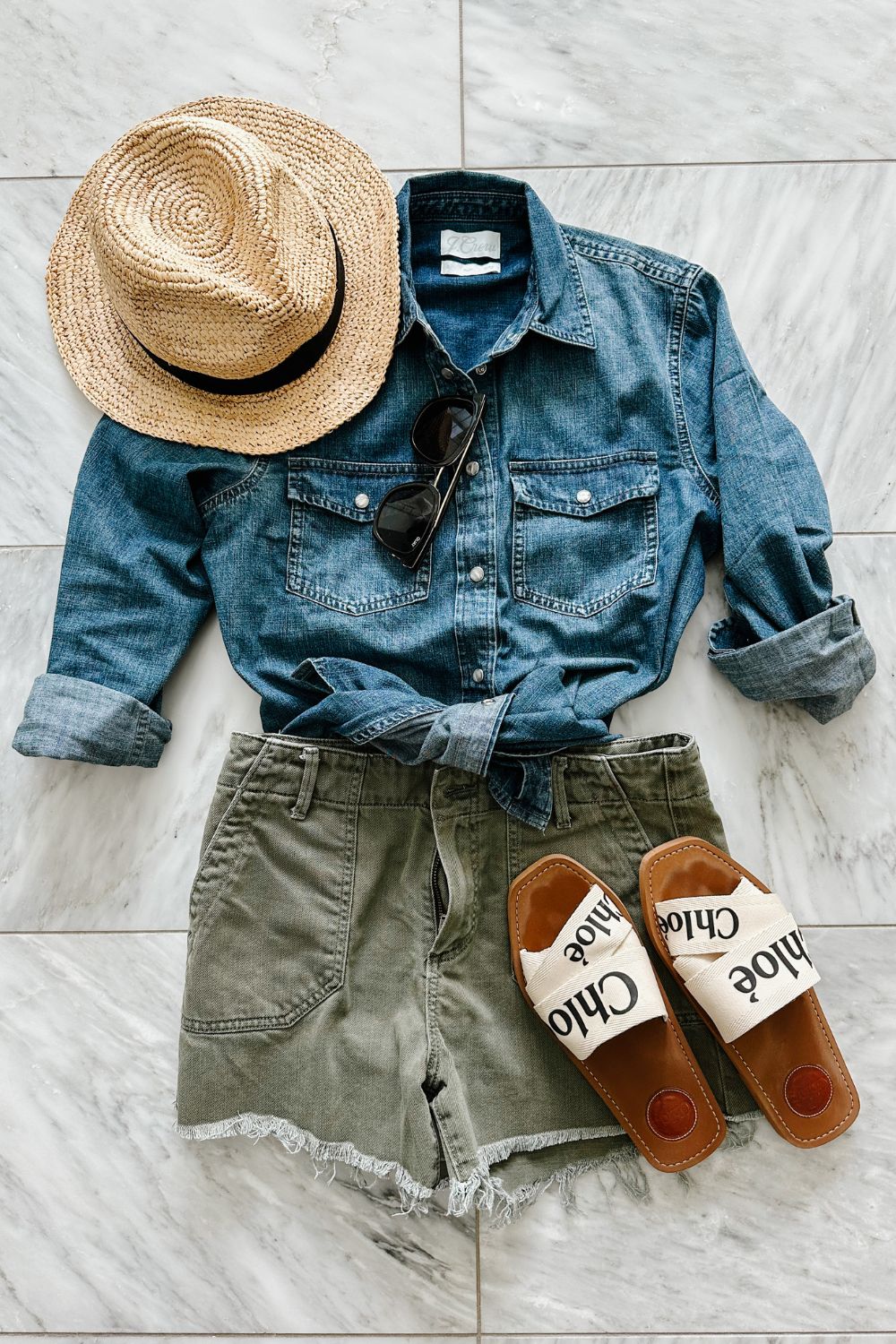 A chambray shirt and olive shorts styled with sandals, sunglasses, and a hat 